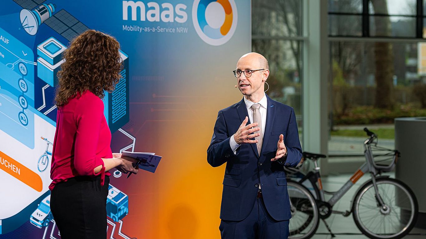 Eine Frau und ein Mann stehen vor einer großen Pappwand, auf der MaaS Mobility-as-a-Service NRW steht.